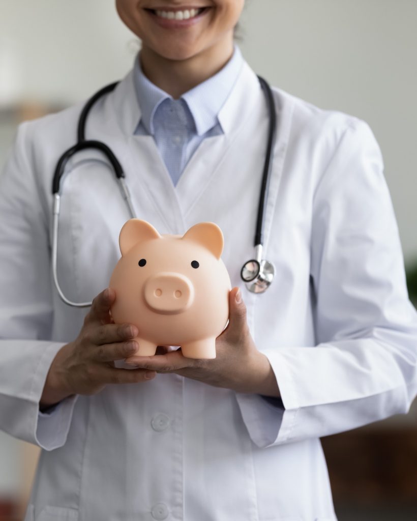 Close up happy young Indian female GP doctor holding piggy bank in hands, advertising collecting costs for medical insurance or charity donation help in clinic office, healthcare and finances concept.
